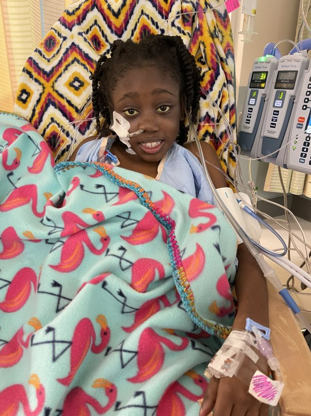 Bra'laya lies in a hospital bed, wrapped in a flamingo blanket, connected to medical equipment.